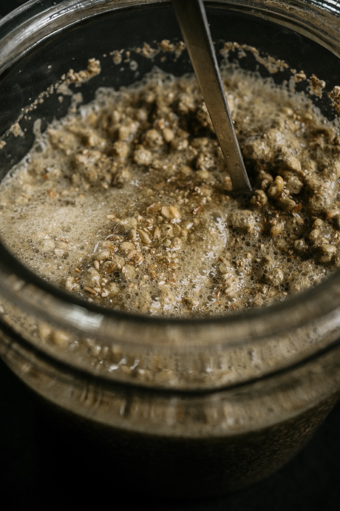 Blick von oben in ein Einmachglas mit dunkler, sprudelnder Fermentmasse, ein Löffel steckt darin
